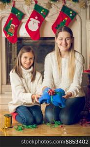 Happy young mother and daughter sitting on floor at fireplace and packing sweater for Christmas