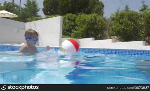 Happy young mother and little son wearing goggles having fun with ball in the swimming pool by the summer house. Holidays at the villa
