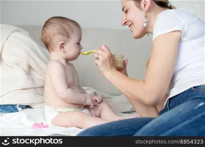 Happy young mother feeding her baby on bed with a spoon