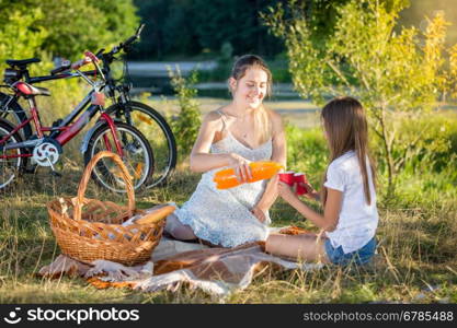 Happy young mother having picnic by the river with daughter. Mother pouring orange juice in daughter's cup