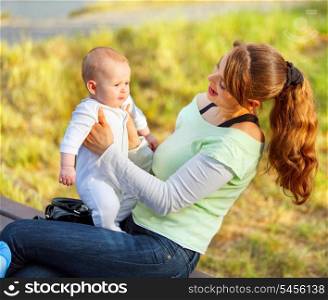 Happy young mother sitting on bench in park and holding baby in hands&#xA;