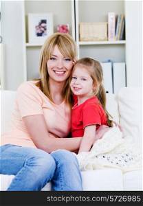 Happy young mother sitting on the sofa and embracing her loving little daughter - indoors