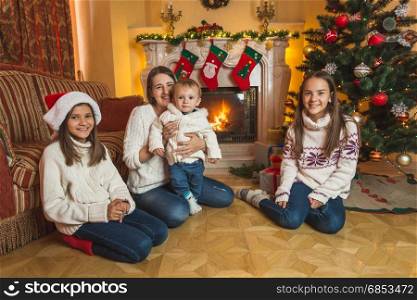 Happy young mother sitting with children on the floor at fireplace. Decorated Christmas tree on background.