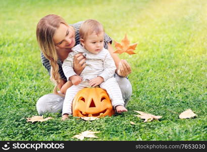 Happy young mother teaching her precious little son about autumnal leaves, cheerful baby sitting on the pumpkin decorative toy, Halloween holiday celebration
