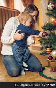 Happy young mother with her 10 months old baby boy decorating Christmas tree at living rom