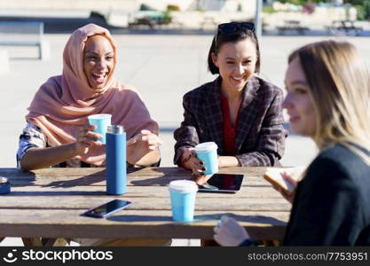 Happy young multiethnic female friends, in casual clothes, smiling while resting in outdoor cafe and drinking coffee from take-away cups. Happy diverse women having coffee break and chatting outdoors