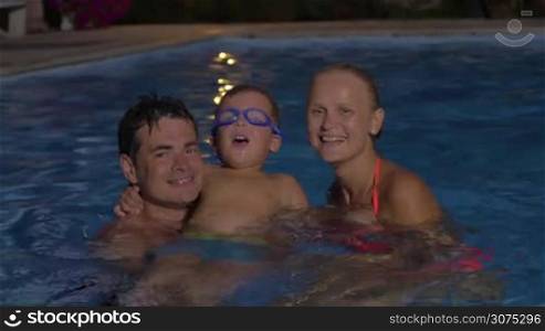Happy young parents and little son looking to the camera with smile while bathing in the pool at night