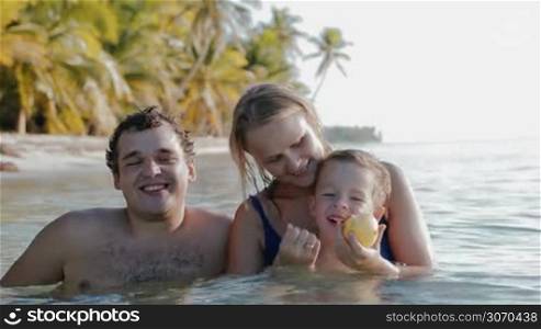 Happy young parents and little son relaxing in sea water close to shore with palms. Mother and son eating apple