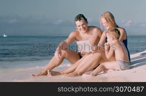 Happy young parents and little son sitting on the beach and watching video or photos on tablet computer. Water washing their feet