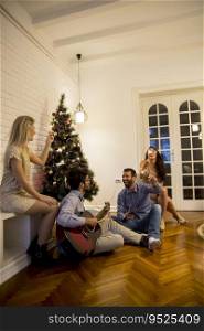Happy young people celebrating Christmas and New Year by fir-tree at home and playing acoustic guitar