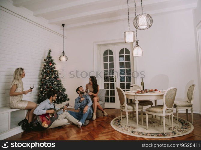 Happy young people celebrating Christmas and New Year by fir-tree at home and playing acoustic guitar