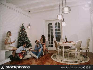 Happy young people celebrating Christmas and New Year by fir-tree at home and playing acoustic guitar