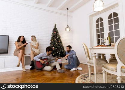 Happy young people celebrating Christmas and New Year by fir-tree at home and playing acoustic guitar