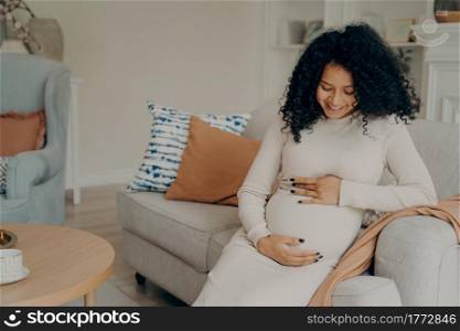 Happy young pregnant afro american lady in white dress with long curly hair looking at her belly with gentle smile, taking little break from housework by resting on couch and drinking cup of green tea. Pregnant afro american lady taking break on couch at home, talking with future baby