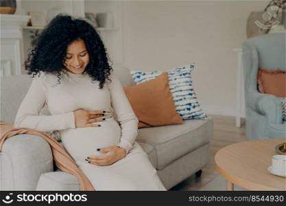 Happy young pregnant afro american lady in white dress with long curly hair looking at her belly with gentle smile, taking little break from housework by resting on couch and drinking cup of green tea. Pregnant afro american lady taking break on couch at home, talking with future baby