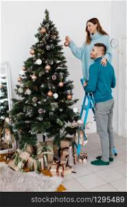 Happy young pregnant couple decorating Christmas tree. Man and woman in stylish clothes celebrate Christmas holiday at home. Smiling Man and Woman together Celebrating Christmas or New Year.