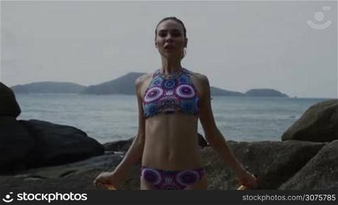 Happy young pretty blonde female wearing colorful bikini exercising using jumping rope between the rocks near the sea during summer day - video in slow motion