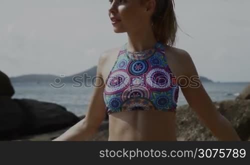Happy young pretty blonde female wearing colorful bikini exercising using jumping rope between the rocks near the sea during summer day - video in slow motion