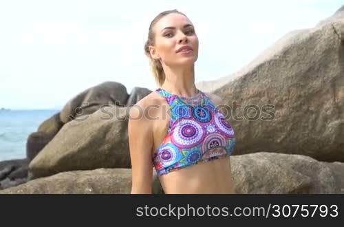 Happy young pretty blonde female wearing colorful bikini exercising using jumping rope between the rocks near the sea during summer day