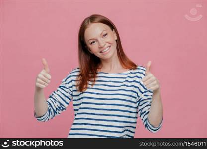 Happy young pretty European lady give double thumb up, likes somebody idea or gives approval, dressed in striped outfit, isolated over pink background. People, body language, gesturig concept
