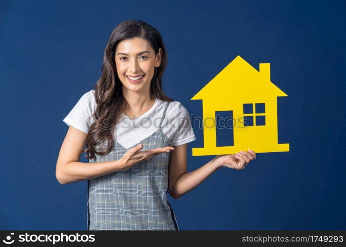 Happy young smiling Asian woman holding and Presenting yellow color house mockup paper on blue color background, thinking for investment, sales and insurance, building and property concept 