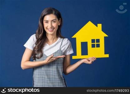 Happy young smiling Asian woman holding and Presenting yellow color house mockup paper on blue color background, thinking for investment, sales and insurance, building and property concept 