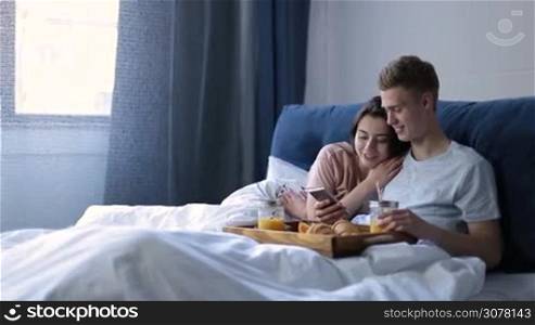 Happy young smiling couple having romantic breakfast in bed in the morning. Handsome hipster surfing the net with smartphone while his pretty girlfriend cuddling him. Cheerful couple in love relaxing in bed at home.