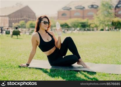 Happy young sporty European woman has rest after training poses on fitness mat with bare feet drinks water dressed in activewear sunglasses leads healthy lifestyle. People workout hydration concept