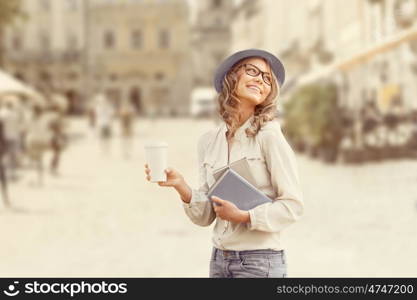 Happy young student with a coffee-to-go, holding books for reading and studying against urban city background.