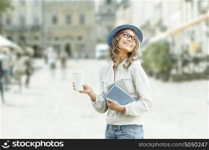 Happy young student with a coffee-to-go, holding books for reading and studying against urban city background.