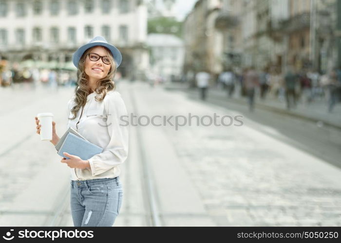 Happy young student with a coffee-to-go, holding books for reading and studying against urban city background.