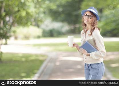 Happy young student with a coffee-to-go, walking in a summer park and holding books for reading and studying.