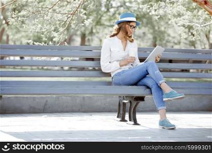 Happy young student with a tablet and a disposable coffee cup sitting on the bench and reading in a summer park.