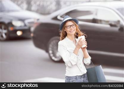 Happy young trendy student holding a coffee-to-go cup and standing at the crosswalk in an urban city.