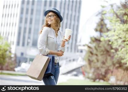 Happy young trendy woman drinking take away coffee and walking with shopping bags after shopping in an urban city.