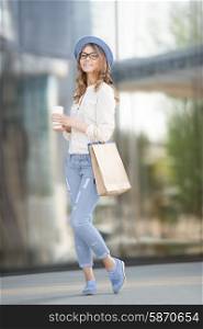 Happy young trendy woman drinking take away coffee, standing on the stairs with shopping bags, and looking aside with smile against urban city background.