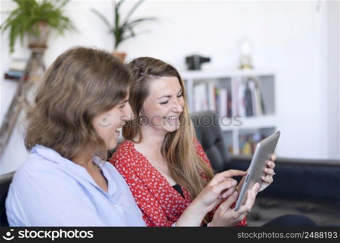 Happy young two blonde women sitting in the couch looking at a tablet