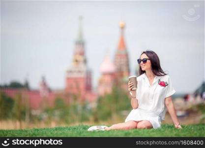 Happy young urban woman drinking coffee outdoors in big city relaxing on the grass. Portrait of smiling girl with drinks in hands.. Happy young urban woman drinking coffee in european city.