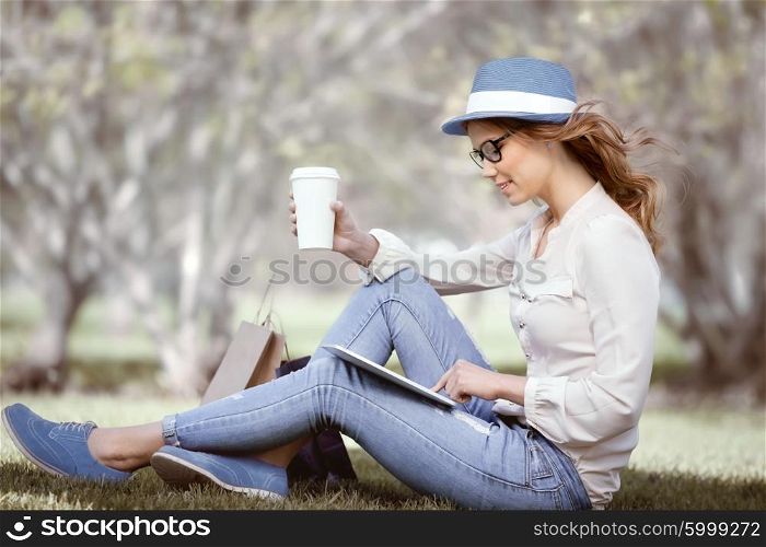 Happy young woman a disposable coffee cup sitting on the grass and using touch pad on her tablet in a summer park.