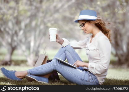 Happy young woman a disposable coffee cup sitting on the grass and using touch pad on her tablet in a summer park.