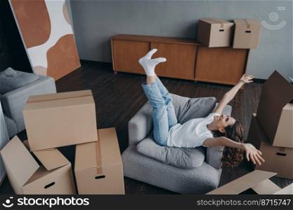 Happy young woman celebrating relocation to first new home, having fun packing, raising hands sitting in armchair on day of moving, excited girl do yes gesture rejoicing surrounded by cardboard boxes.. Excited woman celebrates relocation to new home, rejoicing surrounded by carton boxes on moving day