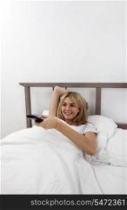 Happy young woman changing channels with remote control in bed