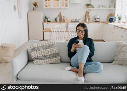 Happy young woman chatting online in social networks on mobile phone, sitting on couch at home. Female reading message on smartphone, getting good news shopping on internet stores.. Happy woman chatting in social networks on smartphone, shopping online, sitting on couch at home