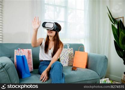 Happy young woman customer wearing 3d VR glasses virtual reality headset with shopping bags around she touching and pointing on air to online shopping in living room at home, digital cyberspace