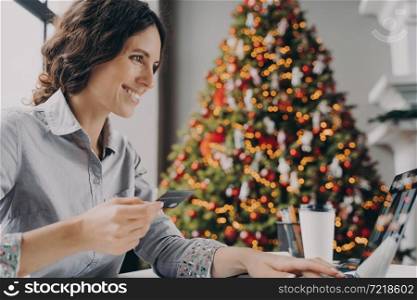 Happy young woman doing online shopping on laptop computer at home during Winter holidays sales, smiling hispanic female with credit card making payment while sitting at table near xmas tree. Happy young woman doing online shopping on laptop computer at home during Winter holidays sales