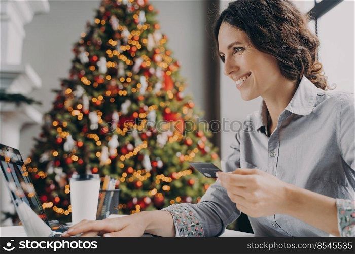 Happy young woman doing online shopping on laptop computer at home during Winter holidays sales, smiling hispanic female with credit card making payment while sitting at table near xmas tree. Happy young woman doing online shopping on laptop computer at home during Winter holidays sales