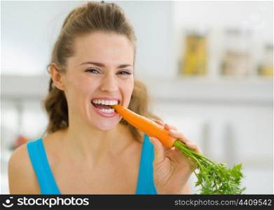 Happy young woman eating carrot in kitchen