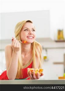 Happy young woman eating fresh fruits salad in kitchen