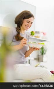 Happy young woman eating fresh salad in modern kitchen