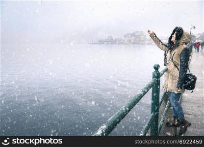 Happy young woman enjoying the first snowfall, reaching to catch snowflakes, on the Hallstatter See lakeshore, in the Hallstatt town, in Austria.
